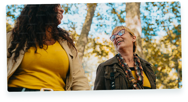 Two women smiling at each other outdoors, one with long dark curly hair, the other with short blonde hair and glasses, with trees and sunshine in the background.