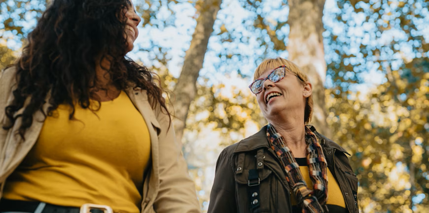 Two women smiling at each other outdoors, one with long dark curly hair, the other with short blonde hair and glasses, with trees and sunshine in the background.
